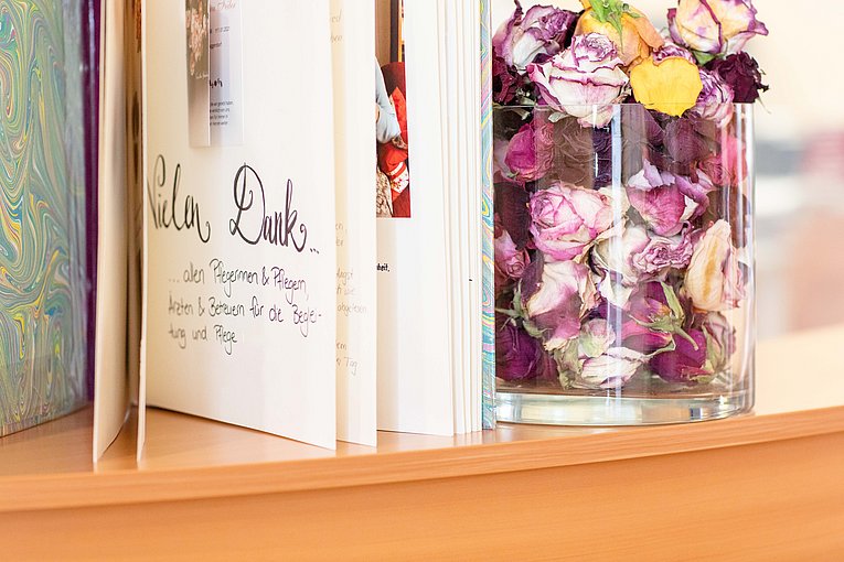 Ein Danke-Buch steht, sodass man ein wenig in einzelne Seiten Einblicken kann an ein großes Glas voller Rosenköpfe gelehnt. 
