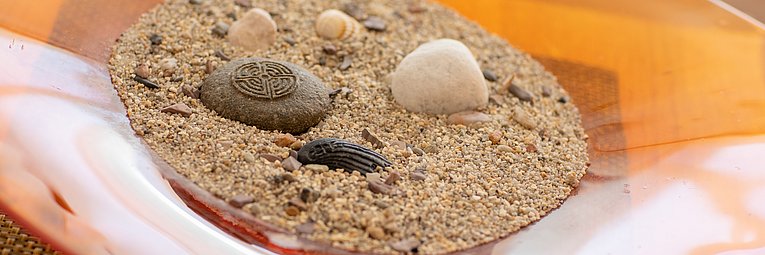In einer flachen orangenen Schale ist grober Sand mit verschiedenen Steinen und Muscheln. 