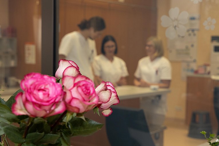 Im Vordergrund sind mehrere Rosen zu sehen. Im Hintergrund besprechen sich drei Pflegekräfte, weiß gekleidet im Stützpunkt der Station.
