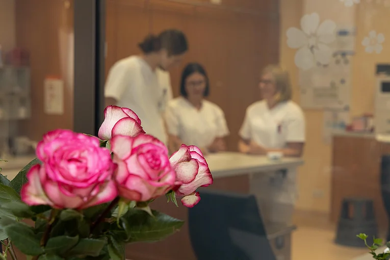 Im Vordergrund sind mehrere Rosen zu sehen. Im Hintergrund besprechen sich drei Pflegekräfte, weiß gekleidet im Stützpunkt der Station. 