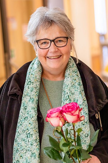 Frau Annette Lechner-Schmidt die evangelische Klinikpfarrerin lächelt in die Kamera. Sie steht auf Station und hält zwei rosa Rosen in der Hand.  