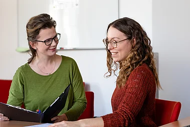 Angehörige.png Zwei Damen sitzen an einem Tisch. Eine davon hält eine Mappe in der Hand. Sie führen eine Unterhaltung mit netten Gesichtsausdruck.